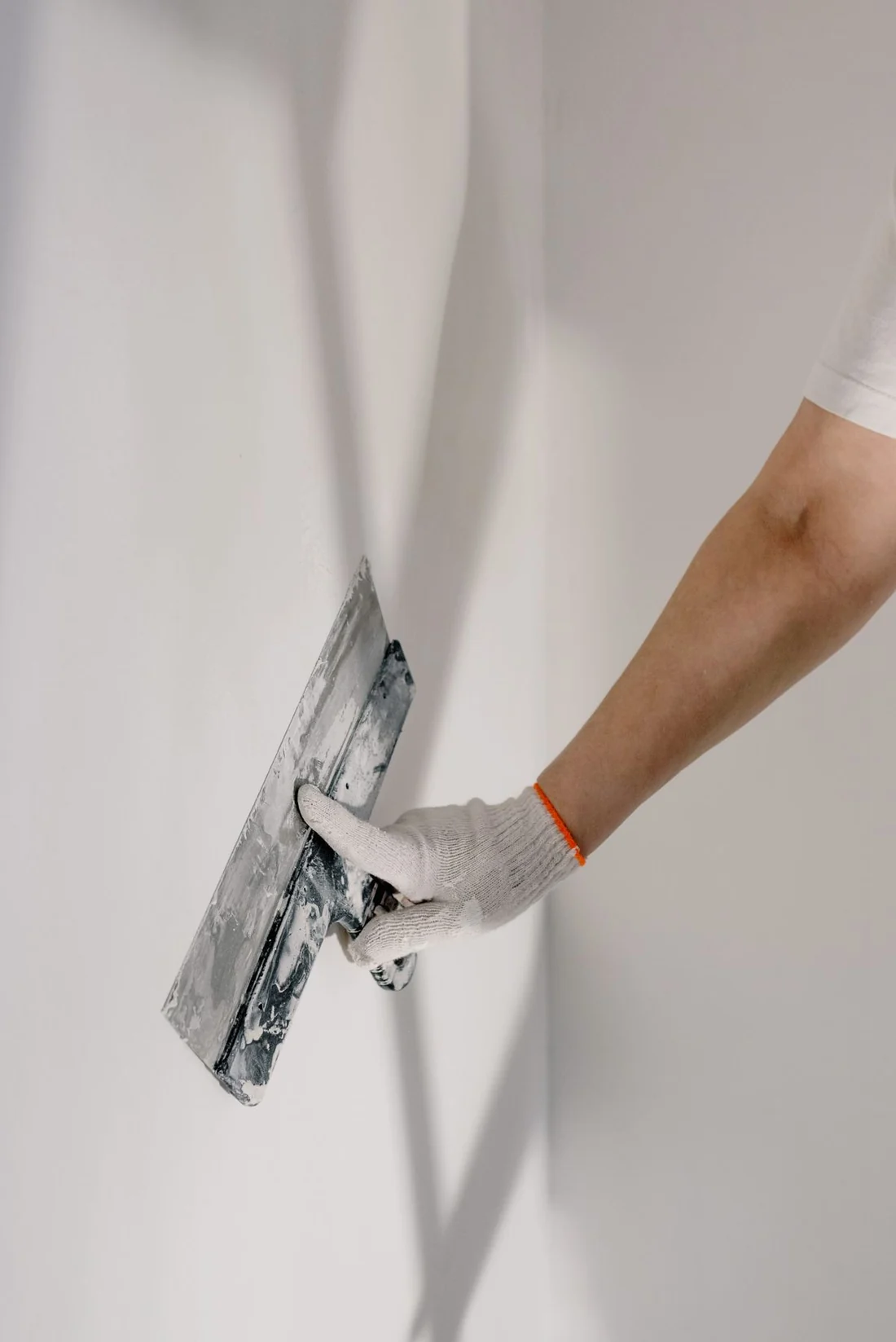 A gloved hand smoothing a freshly skimmed white plaster wall with a steel finishing trowel